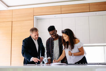 Couple signing a contract documents while standing inside a house with real estate agent. Interracial couple with realtor signing mortgage contract. - JLPSF01441