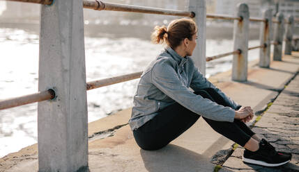Läuferin sitzt nach dem Training an der Strandpromenade. Frau sitzt im Freien und entspannt sich nach dem Lauftraining. - JLPSF01460