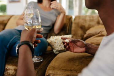 Ein Mann sitzt mit Wein und Popcorn auf dem Sofa, während sich die Frau im Hintergrund entspannt. Ein Paar trinkt zu Hause Wein und isst Popcorn. - JLPSF01471