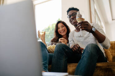 Ein gemischtrassiges Paar sitzt mit einem Glas Wein auf dem Sofa und isst Popcorn, während es einen Film auf dem Laptop im Wohnzimmer anschaut. - JLPSF01472