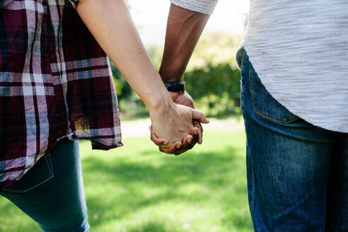 Nahaufnahme eines Paares, das sich an den Händen hält und im Freien spazieren geht. Rückansicht eines Mannes und einer Frau, die sich gegenseitig an den Händen halten, während sie in einem Park spazieren gehen. - JLPSF01473