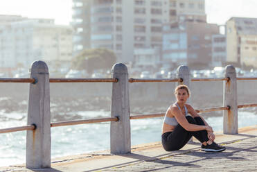 Female runner taking break after fitness training outdoors by the sea. Woman relaxing after morning run on road by the sea. - JLPSF01475