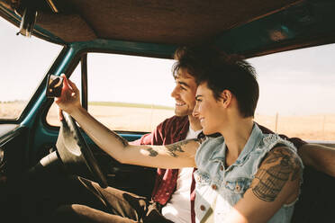 Fröhliches junges Paar auf einer Autoreise, das die Fahrt genießt. Mann und Frau machen ein Selfie im Auto sitzend. - JLPSF01501