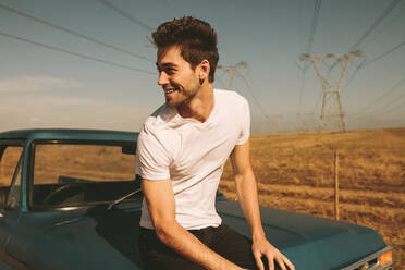 Man sitting on bonnet of a car parked during a road trip to country side. Man enjoying a road trip in country side. - JLPSF01502