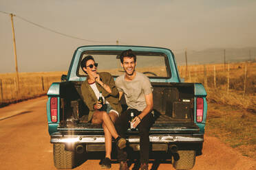 Smiling couple sitting in the back of their pickup truck enjoying the road trip in country side. Man and woman holding a bottle of drink enjoying the road trip. - JLPSF01509