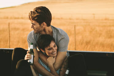 Couple sitting in the back of their pickup truck enjoying the road trip in country side. Man and woman hold each other while enjoying a bottle of drink during a road trip. - JLPSF01510