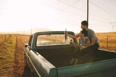 Cheerful couple sitting in the back of their pickup truck enjoying the road trip in country side. Couple taking a selfie using mobile phone sitting at the back of their pick up truck. - JLPSF01512