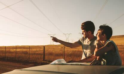 Couple using a map for navigation while on a road trip in country side. Man pointing in the direction of the road while using a map to navigate. - JLPSF01513