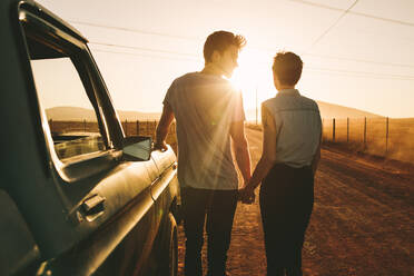 Romantic couple standing holding hands beside their pickup truck during a road trip in country side. Rear view of couple holding hands and standing on a mud track on highway. - JLPSF01516