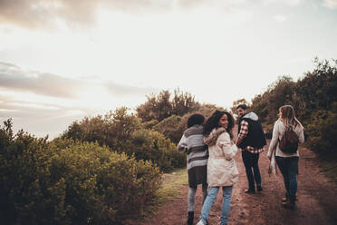 Freunde wandern in der Natur. Eine Gruppe von Männern und Frauen geht auf einer Landstraße spazieren. Die Frau dreht sich um und schaut in die Kamera. - JLPSF01538