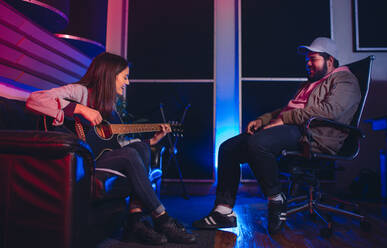 Frau spielt Gitarre und singt, Mann sitzt auf einem Stuhl und hört zu. Musiker komponieren ein Lied auf der Gitarre im Aufnahmestudio. - JLPSF01539