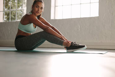 Fit young female sitting on exercise mat in gym. Healthy woman taking break after workout in gym. - JLPSF01563