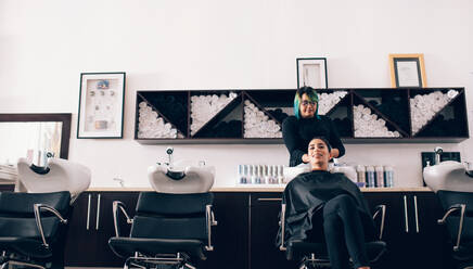 Woman sitting on salon chair while a hair stylist washing her hair. Hairdresser wiping the hair dry using a towel in the wash tub. - JLPSF01604