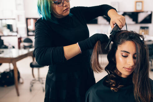 Eine Frau, die sich im Salon frisieren lässt. Ein Friseur verwandelt glattes Haar mit einem Lockenstab in lockiges Haar. - JLPSF01614