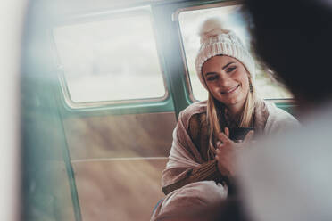 Frau, die mit einem Freund in einem Lieferwagen unterwegs ist. Kaukasische Frau in warmer Kleidung sitzt auf dem Rücksitz des Lieferwagens, hält einen Kaffee in der Hand und lächelt. - JLPSF01621