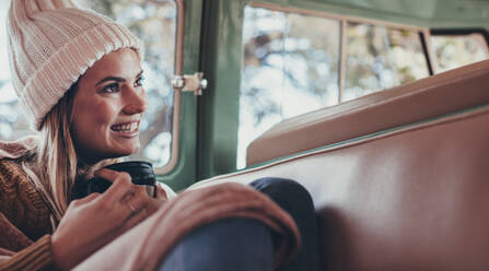 Lächelnde Frau in warmer Kleidung sitzt mit einer Tasse Kaffee auf dem Rücksitz eines Lieferwagens. - JLPSF01622