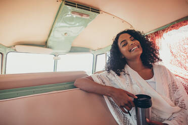 Junge Frau sitzt mit Kaffee auf dem Rücksitz eines Vans und lächelt. Afrikanische Frau genießt die Fahrt. - JLPSF01625