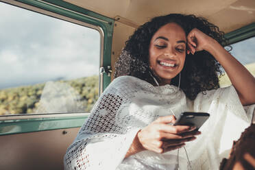 Lächelnde Frau, die auf der Rückbank des Autos sitzt und mit ihrem Handy Musik hört. - JLPSF01649