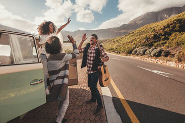 Eine Gruppe von Männern und Frauen auf einem Roadtrip, die neben dem Van stehen und sich ein High Five geben. Fröhliche Freunde, die sich im Urlaub amüsieren. - JLPSF01660