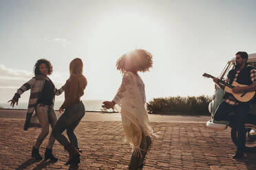 Eine Gruppe von Freunden auf einem Roadtrip tanzt und hat Spaß. Ein Mann spielt Gitarre und Frauen tanzen auf der Straße. - JLPSF01673
