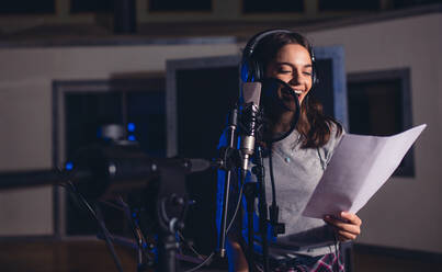 Smiling female singer with microphone and reading lyrics. Woman recording a song in music studio. Female playback singer recording her new album. - JLPSF01674