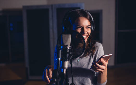 Lächelnde Playback-Sängerin mit Mikrofon und Mobiltelefon im Aufnahmestudio. Frau nimmt einen Song für ihr Album im Musikstudio auf. - JLPSF01678
