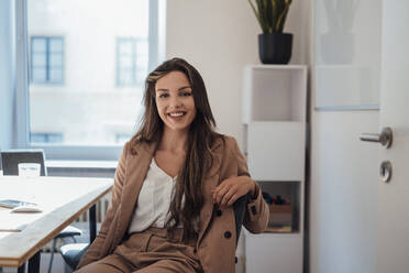 Lächelnde junge Frau mit langen Haaren, die in einem Büro zu Hause sitzt - JOSEF13388