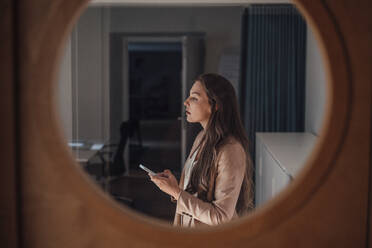 Nachdenkliche Geschäftsfrau mit langen Haaren, die ein Smartphone hält, gesehen durch ein Glas - JOSEF13394