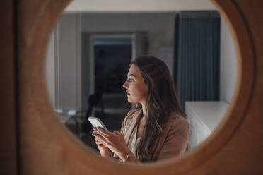 Thoughtful businesswoman with mobile phone seen through glass - JOSEF13395