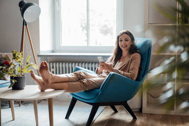 Happy businesswoman with smart phone sitting on chair at home - JOSEF13396