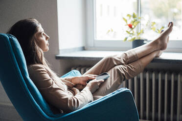 Thoughtful young businesswoman sitting on chair with feet up at home - JOSEF13398