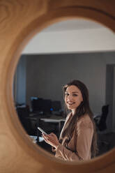 Smiling young businesswoman with smart phone seen through glass - JOSEF13400