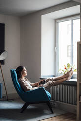 Thoughtful businesswoman sitting on chair with feet up at home - JOSEF13404