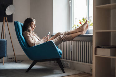 Geschäftsfrau mit hochgelegten Füßen und Smartphone auf einem Stuhl zu Hause - JOSEF13412