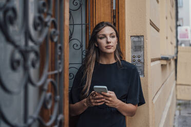 Nachdenkliche junge Frau mit Smartphone an der Tür stehend - JOSEF13413