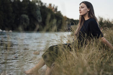 Nachdenkliche junge Frau mit langen Haaren sitzt am See - JOSEF13417