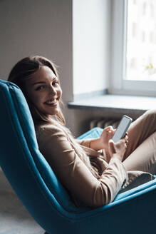 Glückliche Geschäftsfrau auf einem Stuhl sitzend mit Smartphone zu Hause - JOSEF13419
