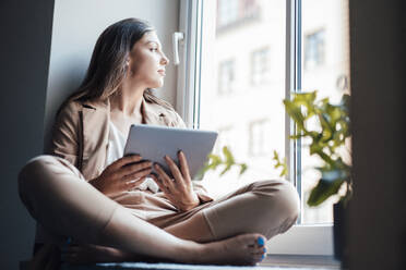 Businesswoman sitting with tablet PC on window sill at home - JOSEF13422