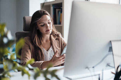 Junge Geschäftsfrau mit Smartphone vor einem Desktop-PC sitzend - JOSEF13425