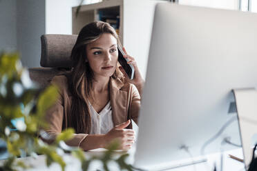 Young businesswoman talking on mobile phone in front of desktop PC - JOSEF13426