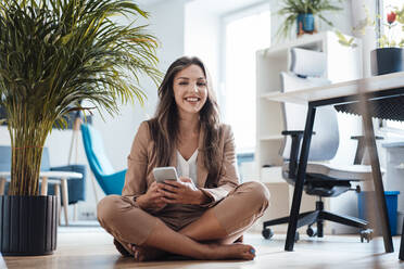 Happy young businesswoman with mobile phone sitting in home office - JOSEF13431