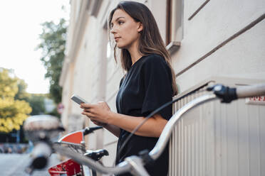 Woman with smart phone standing by bicycle - JOSEF13452