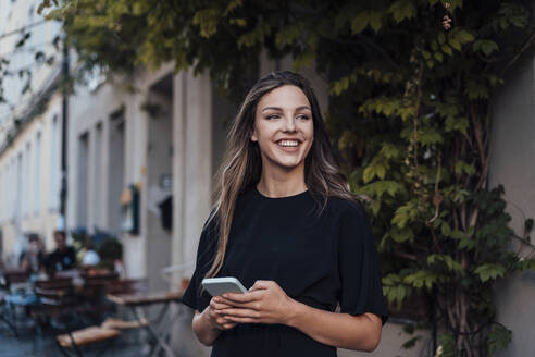 Fröhliche junge Frau mit Smartphone vor einem Straßencafé - JOSEF13455