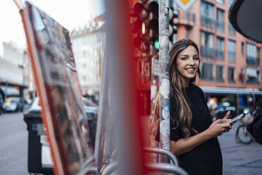 Lächelnde junge Frau, die an einem Pfahl mit einem Mobiltelefon auf der Straße steht - JOSEF13456