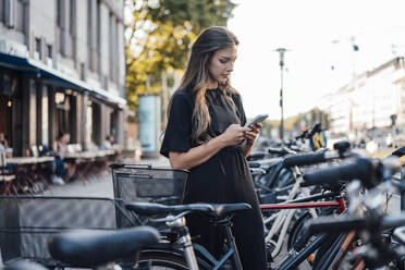 Junge Frau benutzt ein Mobiltelefon an einer Fahrradabstellanlage - JOSEF13457