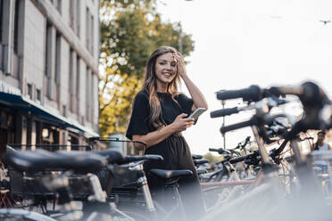 Happy young woman with smart phone standing at bicycle parking station - JOSEF13458