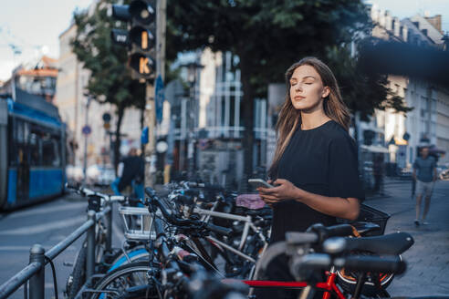 Junge Frau steht mit geschlossenen Augen und hält ihr Smartphone an einer Fahrradabstellanlage - JOSEF13459