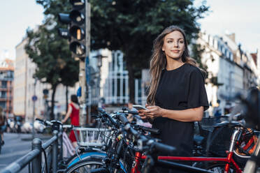 Young woman with smart phone standing at bicycle parking station - JOSEF13460