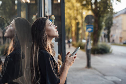 Junge Frau mit Mobiltelefon an einer Glaswand stehend - JOSEF13468