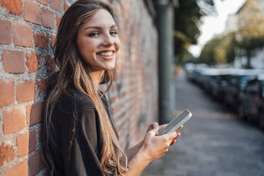 Happy young woman with mobile phone standing by brick wall - JOSEF13473